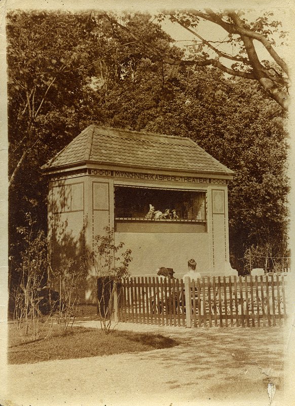 Historische Fotografie: kleiner Theaterpavillon mit Walmdach und Bühnenöffnung hinter Holzzaun, Zuschauer davor