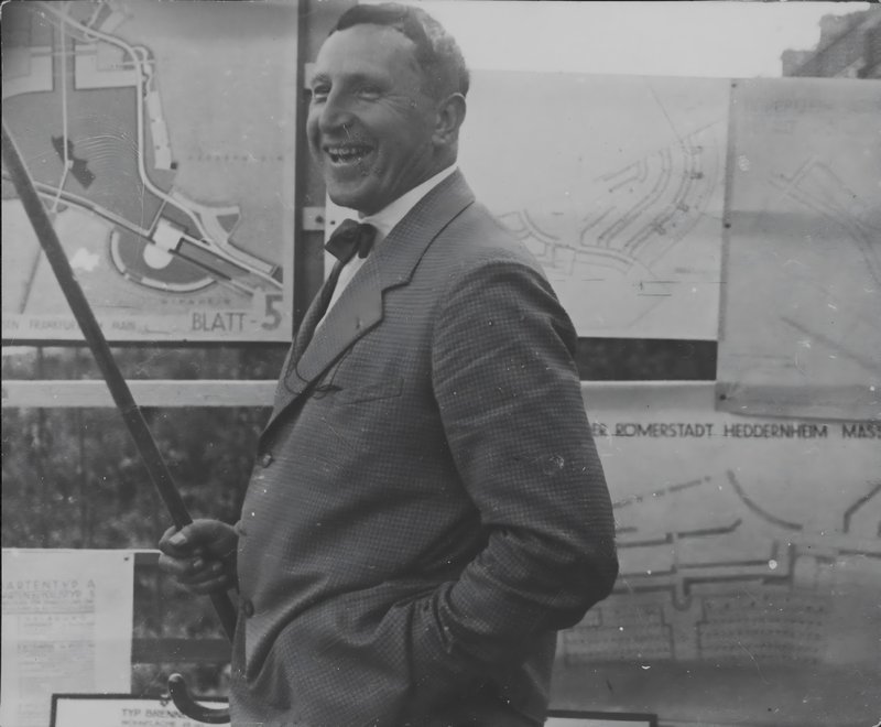 Lachender Ernst May mit Fliege und Zeigestock vor Plantafeln der Siedlung Römerstadt