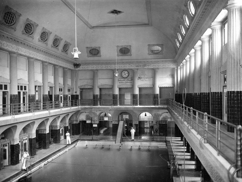 Schwarzweißfoto: Schwimmhalle mit langem Becken, umlaufenden Galerien, Rundfenstern und Uhr