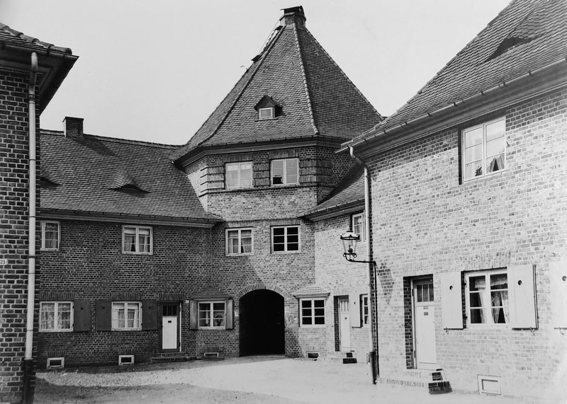 Backstein-Reihenhäuser um einen Hof, Torbau mit Zeltdach und rundbogiger Durchfahrt