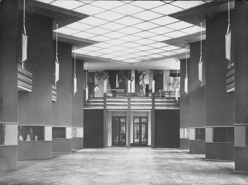 Hohe Pfeilerhalle mit Lichtdecke, dunklen Pfeilern, Galerie und Wandbild an der Stirnseite