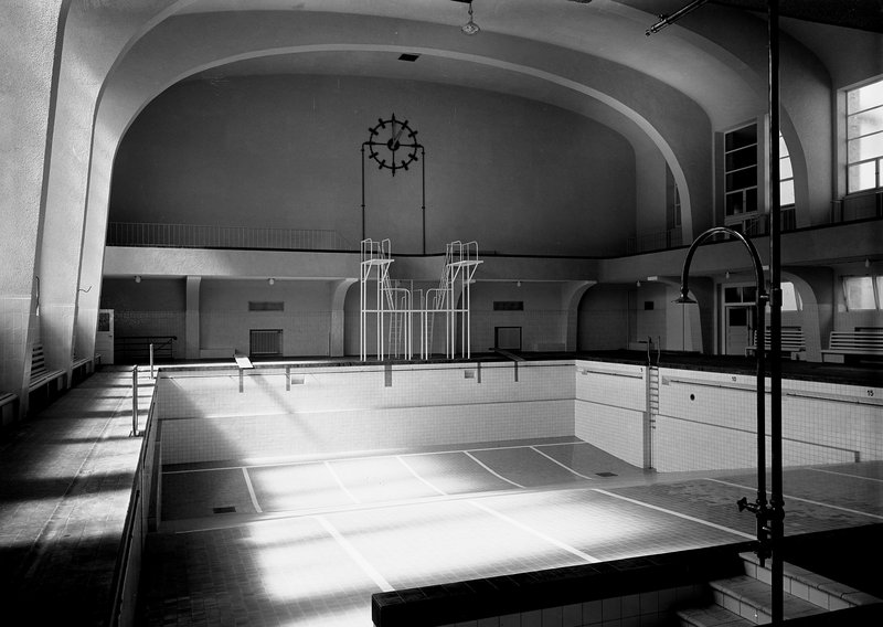 Schwimmhalle mit leerem Becken, Sprungturm und Wanduhr unter gewölbter Decke