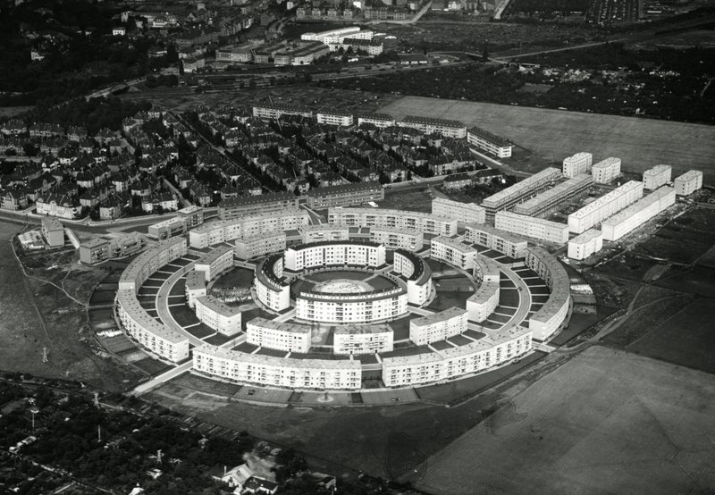 Schrägluftbild der kreisförmigen Siedlung mit drei konzentrischen Gebäuderingen, umgeben von Feldern und Villenvierteln