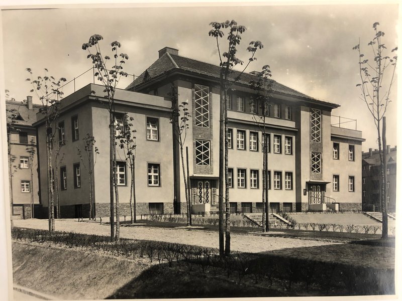 Historisches Foto eines dreigeschossigen Putzbaus mit Walmdach und gitterartigen Fensterbändern, davor junge Bäume