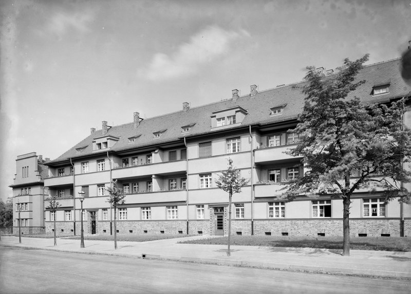Viergeschossiger Wohnblock mit Balkonen und Dachgauben, davor Allee mit jungen Bäumen