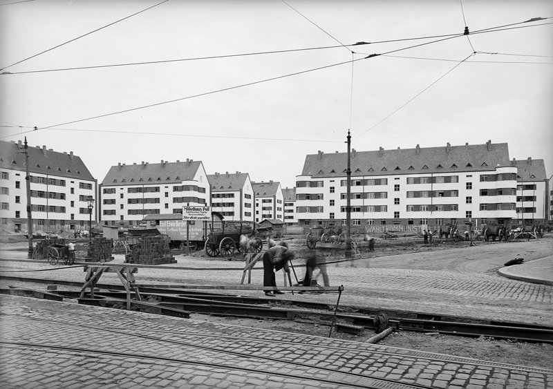 Neue Wohnblöcke mit Satteldach hinter einer Straßenbaustelle mit Arbeitern, Gleisen und Pferdefuhrwerken