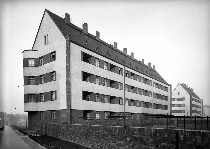 Wohnzeile mit Steildach, abgerundetem Eckerker und Loggien, davor Ziegelmauer und Gehweg