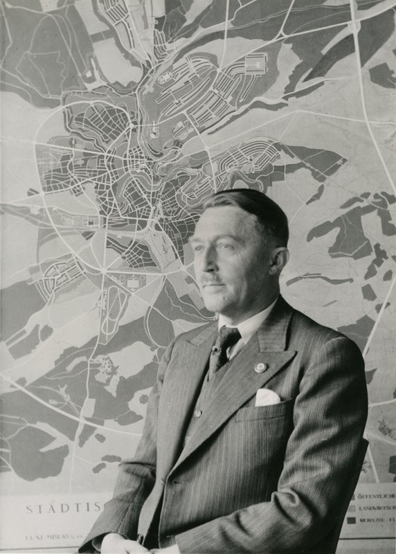 Schwarzweiß-Porträt von Hubert Ritter im Anzug mit Parteiabzeichen vor einem großen Stadtplan