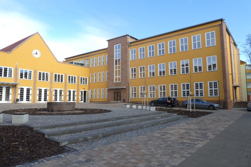 Farbfoto: sanierter gelber Schulbau mit Klinkerlisenen, Giebel mit Uhr und gepflastertem Vorplatz