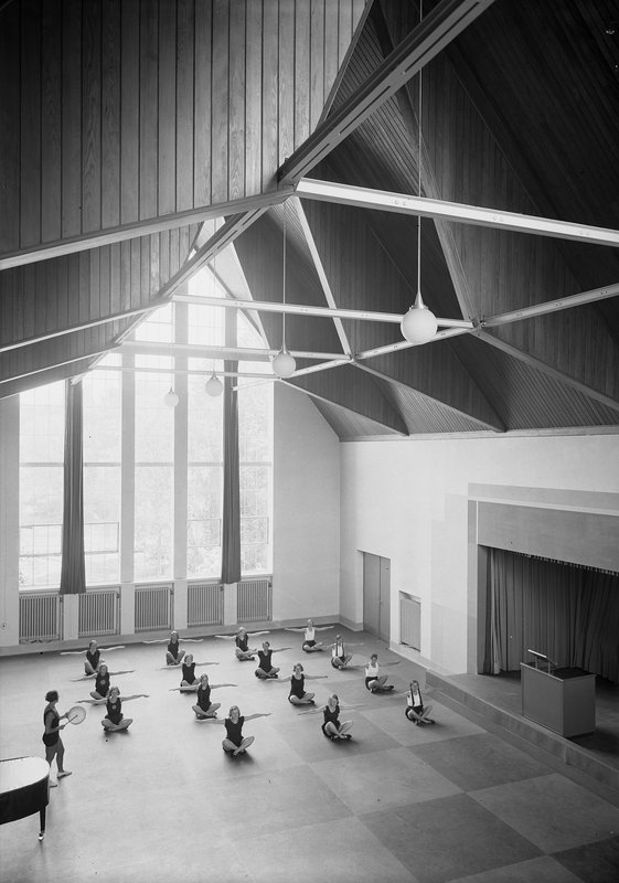Schwarzweißfoto: Mädchen bei Gymnastikübungen in hoher Turnhalle mit Holzdecke, Hängelampen und großer Fensterwand