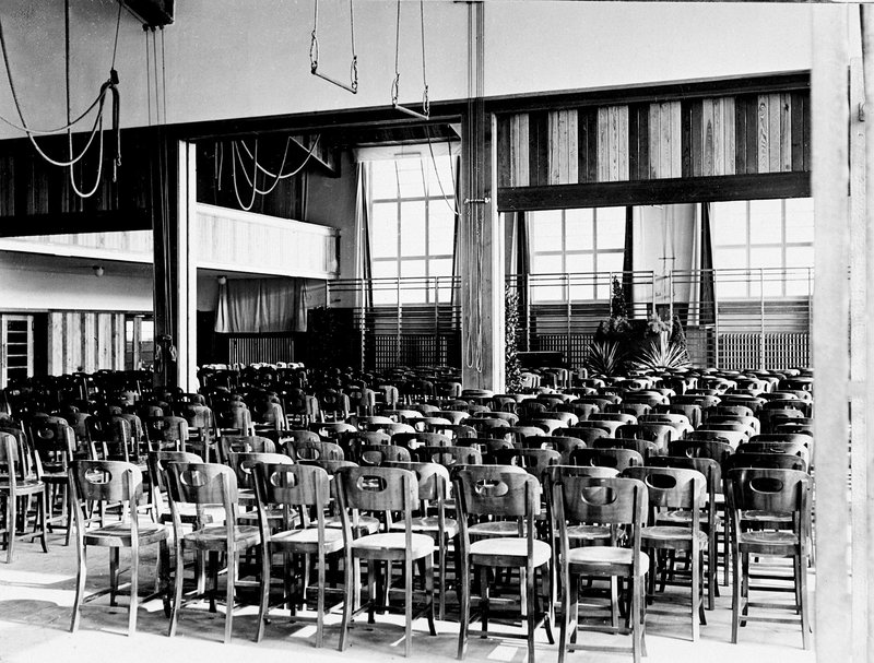 Schwarzweißfoto: bestuhlte Turnhalle mit Ringen an der Decke und großen Fenstern, als Aula genutzt