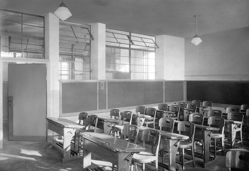 Schwarzweißfoto: Klassenzimmer mit Schulbänken, Tafel und Fensterreihe zur Flurwand