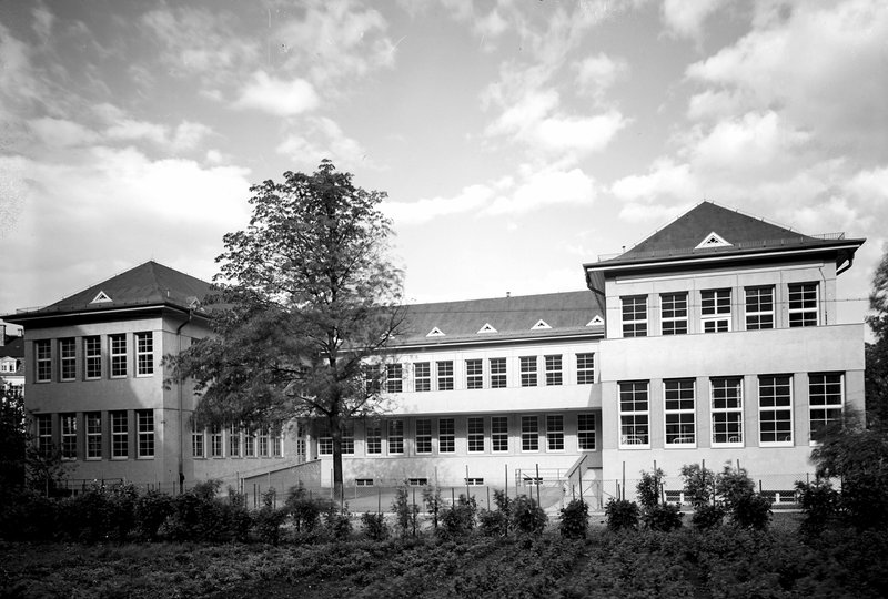 Dreigeschossiger Klinikbau mit Walmdach und großen Fensterreihen, davor Bäume; Schwarz-Weiß-Fotografie