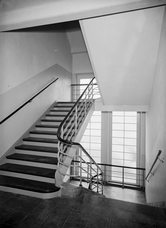 Helles Treppenhaus mit geschwungenem Metallgeländer und hohem Sprossenfenster; Schwarz-Weiß-Fotografie