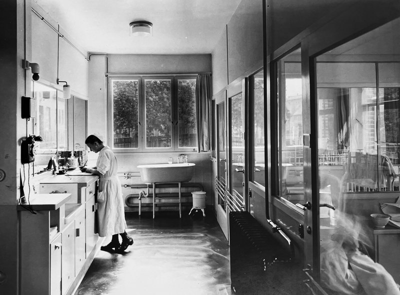 Vorraum mit Waschbecken, Badewanne und Glaswänden, eine Krankenschwester am Waschtisch; Schwarz-Weiß-Foto