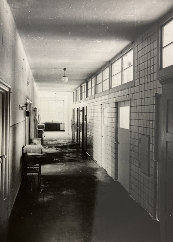 Langer gefliester Klinikkorridor mit Türen links und Fensterband mit Oberlichtern rechts; Schwarz-Weiß-Foto